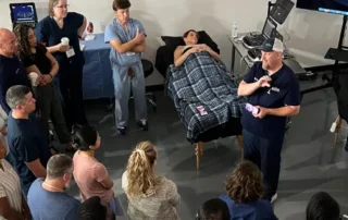 Group of doctors in navy blue scrubs gathered around a patient lying on a hospital bed with arms crossed over their stomach.