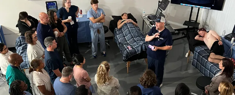 Group of doctors in navy blue scrubs gathered around a patient lying on a hospital bed with arms crossed over their stomach.