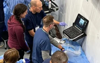 Medical professionals gathered around an ultrasound machine during a hands-on training session. An instructor demonstrates a needle-guided procedure while participants observe closely.