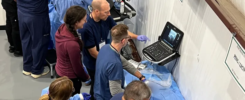 Medical professionals gathered around an ultrasound machine during a hands-on training session. An instructor demonstrates a needle-guided procedure while participants observe closely.